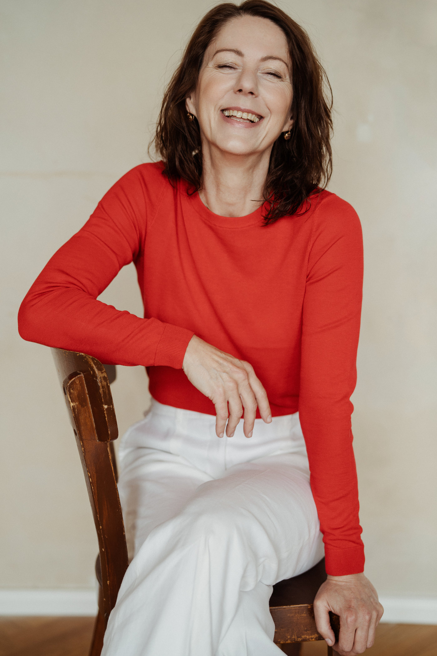 Gabriella Rist, Gründerin der GANESHASHALA® Online Trauma Akademie für Nervensystem und Entwicklungstrauma, lachend auf einem Holzstuhl, im roten Pullover.