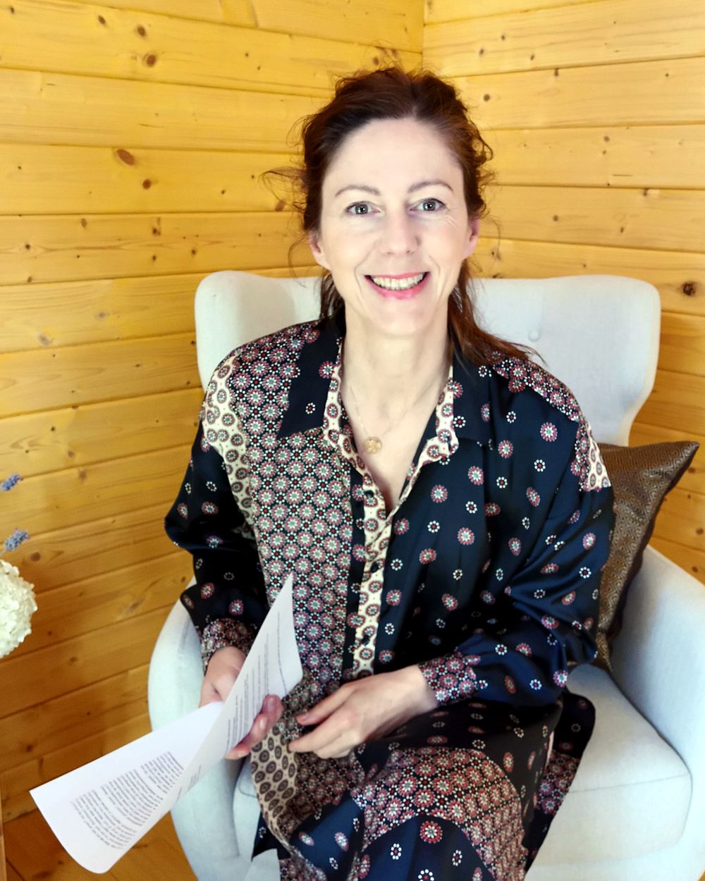 Gabriella Rist sitzt lächelnd in einem hellen Sessel in einem Holzraum, ein Skript in der Hand – Retreat zu Nervensystem und Selbstregulation
