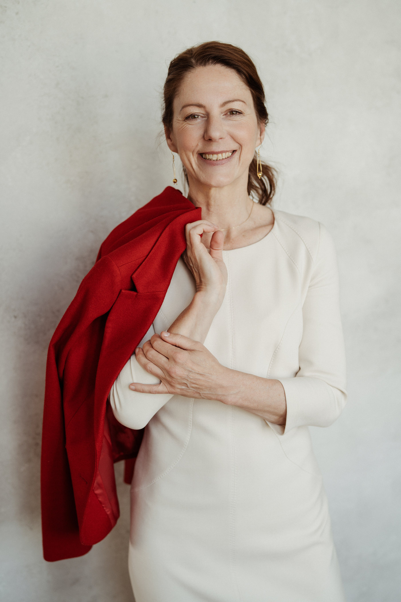 Gabriella Rist im cremefarbenen Kleid, den roten Blazer über der Schulter, die Hand am Herzen – Traumabegleitung mit Blick auf das Nervensystem.
