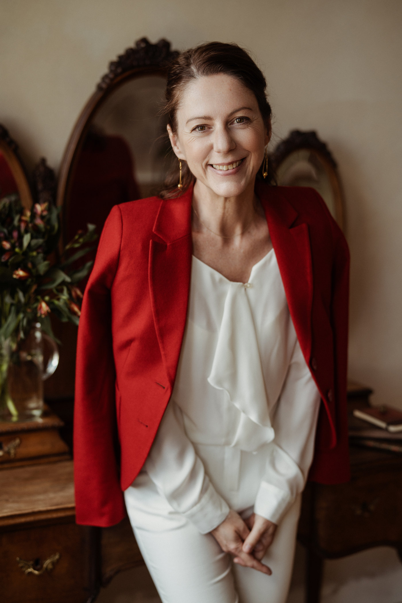Gabriella Rist, Gründerin der GANESHASHALA® Online Trauma Akademie, im roten Blazer vor einem alten Spiegel, lächelnd